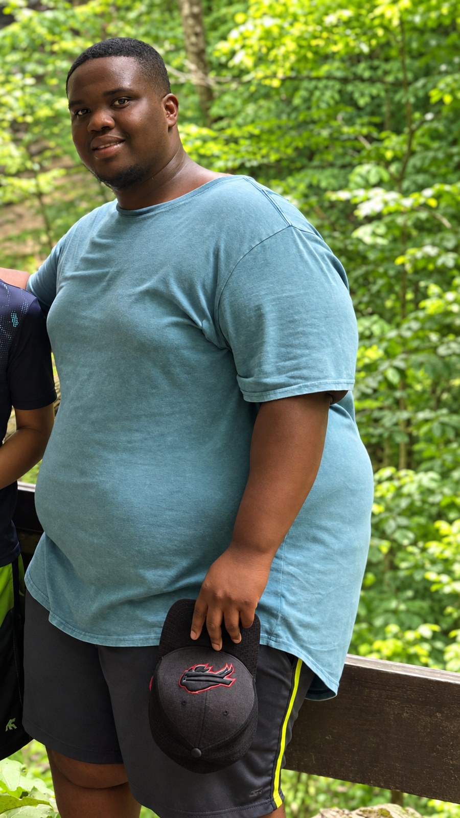 Devrin before, standing outdoors in a teal shirt holding a cap